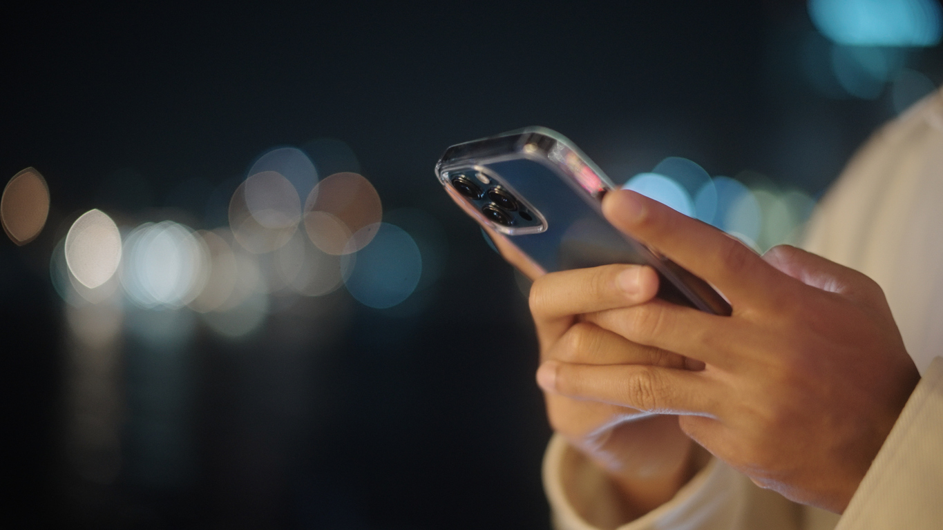 In primo piano si vedono le mani di una persona che utilizza uno smartphone su uno sfondo scuro